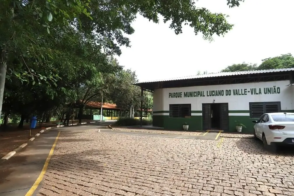 Um prédio branco e verde com a placa “Parque Municipal Luciano do Valle - Vila União” fica ao lado de um estacionamento de paralelepípedos, cercado por árvores, com um carro branco estacionado à direita — um dos parques mais notáveis de Campinas.