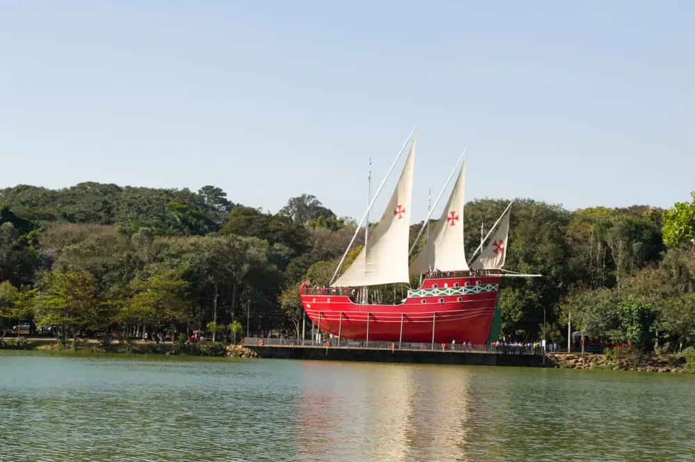 Um grande navio vermelho com velas brancas e cruzes vermelhas está atracado perto da margem de um lago calmo em um dos Parques de Campinas, cercado por árvores e vegetação sob um céu azul e límpido.