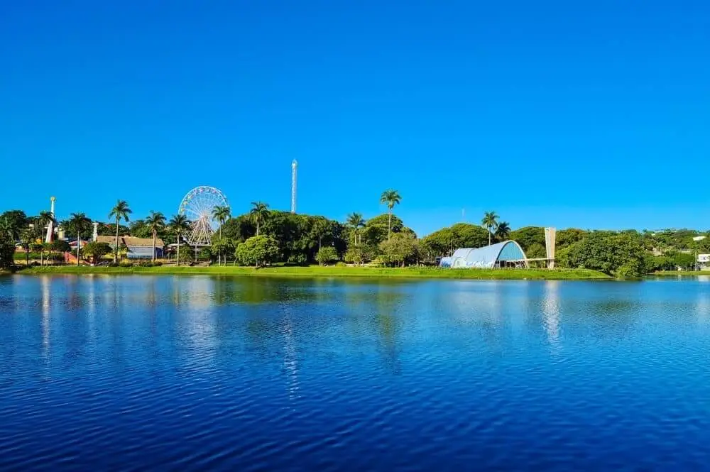 Foto que ilustra matéria sobre ponto turístico de Belo Horizonte mostra uma das margens da Lagoa da Pampulha vista de dentro da água para fora. Em um primeiro plano, aparece parte do espelho d'água. E em terra firme, à direita está a Igreja São Francisco de Assis, ao centro algumas árvores e à esquerda aparece a roda gigante do Parque Guanabara. Ao fundo, o céu está azul