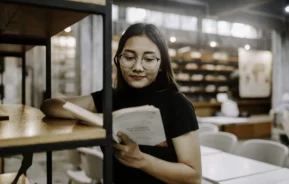 Imagem de uma jovem estudante lendo um livro encostada na prateleira de uma biblioteca para ilustrar matéria sobre as bibliotecas em SP