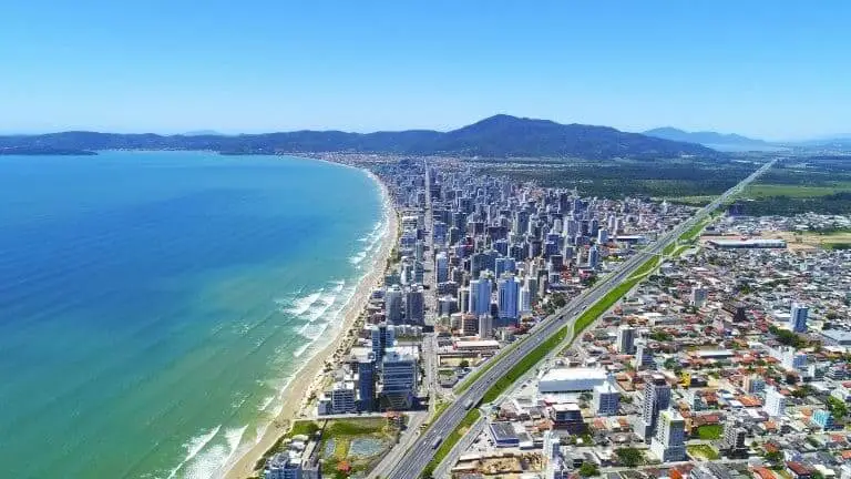 Imagem da vista aérea urbana de Itapema, em Santa Catarina, mostra mar, orla da cidade, prédios, casas, avenidas e vegetação ao fundo em um dia de céu azul para ilustrar matéria sobre as cidades com mais prédios do Brasil