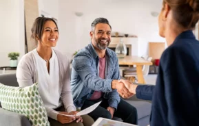 Imagem de um casal formado por um homem e uma mulher sentados no sofá de uma casa em frente a uma agente imobiliária enquanto o homem aperta a mão da profissional para ilustrar matéria sobre como comprar um imóvel que ainda não foi quitado