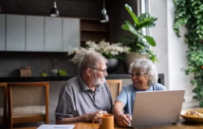 Imagem de um casal de idosos sentados na mesa da cozinha de casa com um notebook na frente enquanto falam sobre as finanças para ilustrar matéria sobre hipoteca reversa