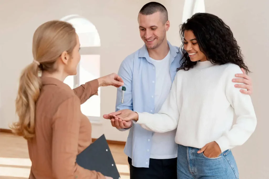 Imagem de uma mulher entregando as chaves de um imóvel para um casal formado por um homem e uma mulher jovens e sorridentes para ilustrar matéria sobre qual é o imposto sobre compra e venda de imóvel