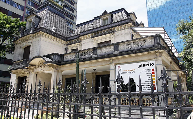 Mansão histórica bege onde fica a Casa das Rosas com telhado de mansarda, pedras ornamentadas e uma cerca na frente. Prédios modernos de vidro a cercam. Uma placa na cerca diz Janeiro com uma escala de cores e texto em português.