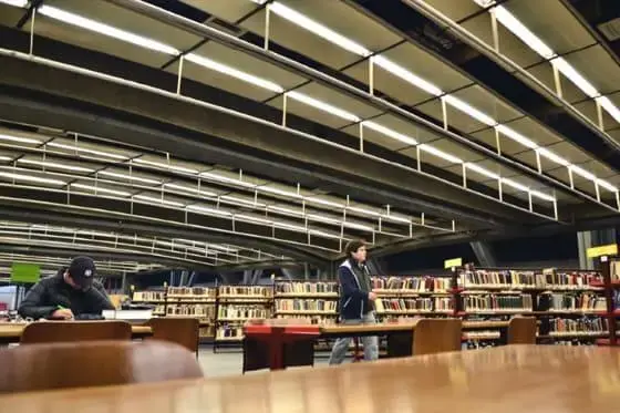 Interior da biblioteca do Centro Cultural de São Paulo mostra fileiras de estantes, mesas e cadeiras. Uma pessoa está sentada lendo ou escrevendo, enquanto outra pessoa passa pelas prateleiras sob luzes de teto modernas e brilhantes