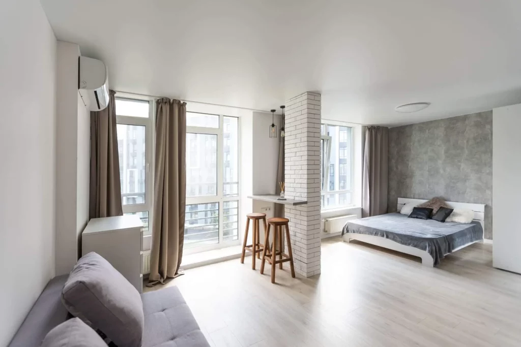 Imagem de uma kitnet moderna e bem iluminada com janelas grandes, piso de madeira clara, um sofá cinza, uma cama com almofadas, uma pequena barra de café da manhã com duas banquetas e cortinas de cores neutras