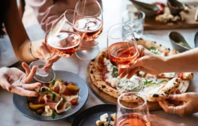 Imagem das mãos de pessoas segurando taças de vinho sobre pratos de comida na mesa de um restaurante para ilustrar matéria sobre melhores restaurantes em Moema