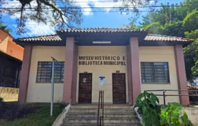 Um prédio bege com acabamento marrom e telhado de telhas, o Museu Histórico Municipal, é um dos notáveis Museus em Guarulhos, com duas portas, janelas gradeadas, degraus, vegetação e árvores sob um céu azul.