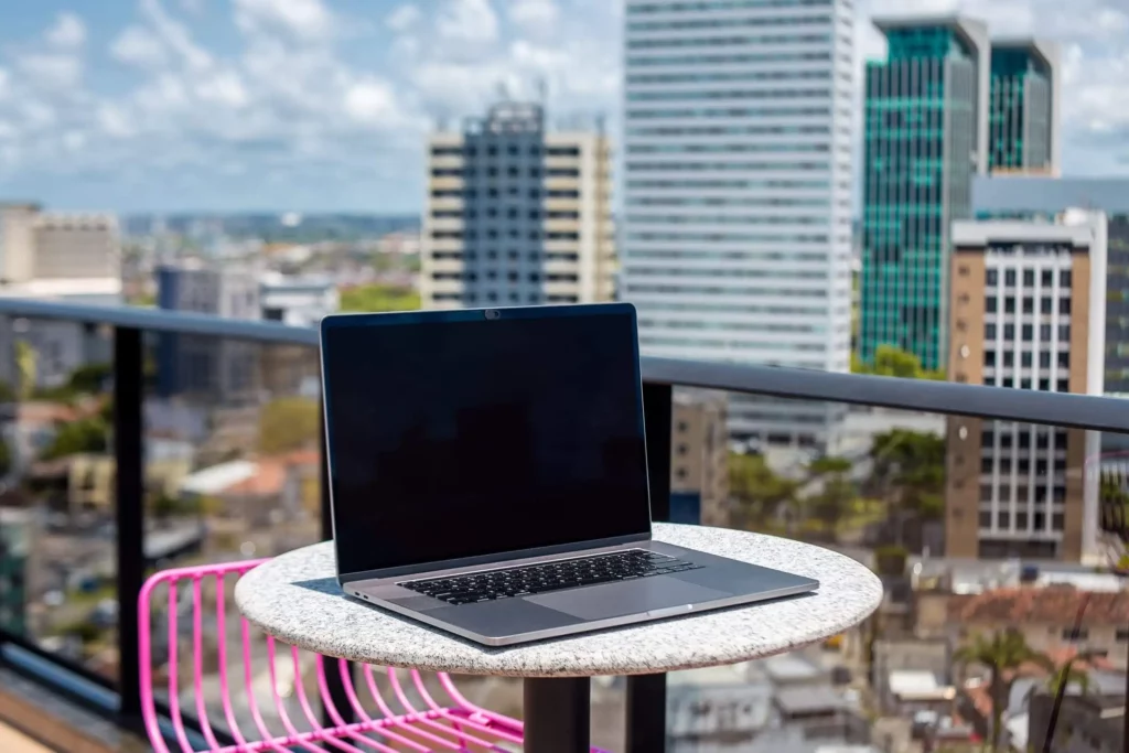 Imagem de um notebook com tela preta está sobre uma mesa redonda ao ar livre em uma varanda, com prédios altos da cidade de Recife, no Brasil, um céu parcialmente nublado ao fundo. Uma cadeira rosa está parcialmente visível ao lado da mesa