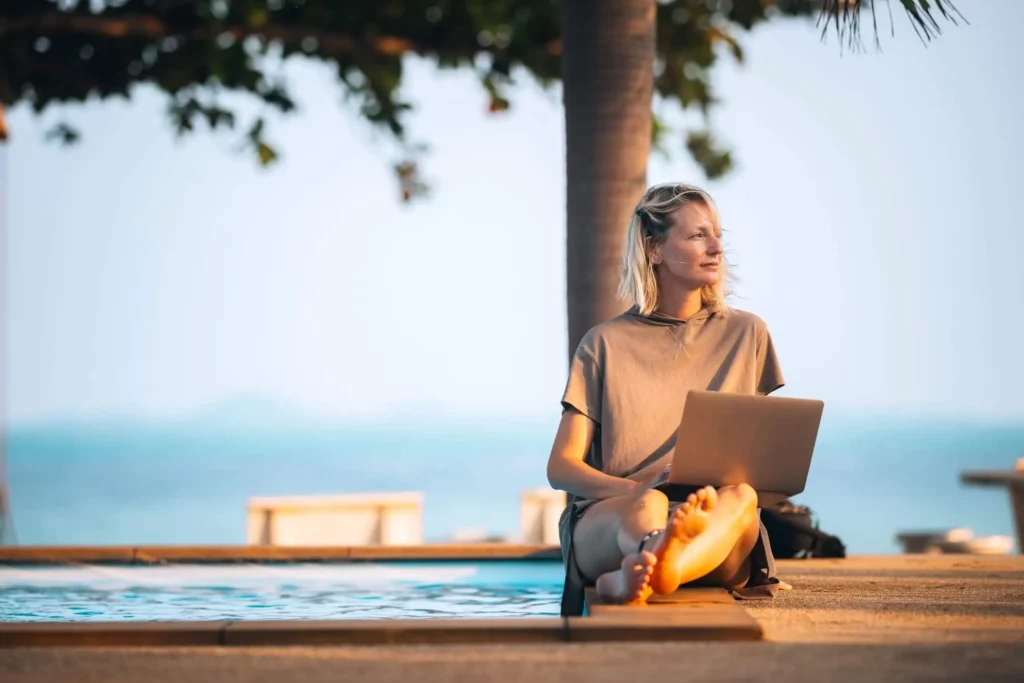 Imagem de uma mulher sentada à beira da piscina com um notebook no colo, descalça e relaxada, olhando para longe. Palmeiras e vistas embaçadas do oceano estão ao fundo, sugerindo um local tropical