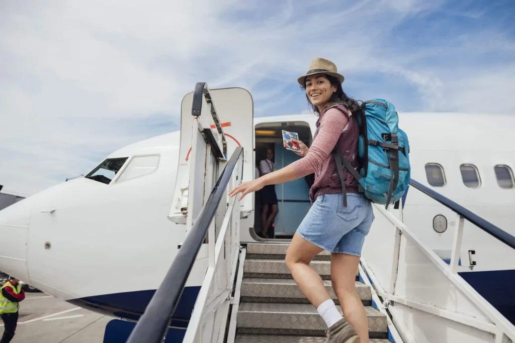 Imagem de um mulher sorridente, usando chapéu e mochila, sobe as escadas para embarcar em um avião, segurando seu passaporte e passagem, com um céu azul ao fundo