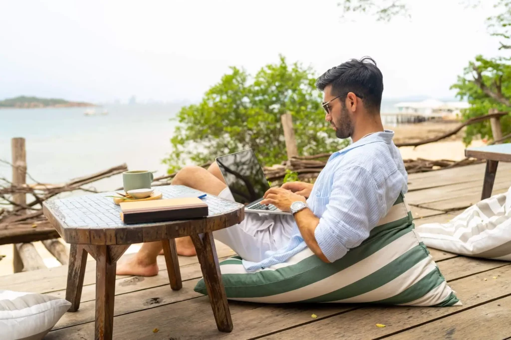 Imagem de um homem sentado em uma almofada listrada em um deck de madeira perto da água, trabalhando em um notebook. Uma mesa próxima a ele contém uma xícara, um pires e livros. Árvores e uma orla calma são visíveis ao fundo
