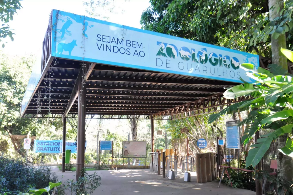 Entrada do Zoológico de Guarulhos com uma grande placa azul onde se lê Sejam Bem-Vindos ao Zoológico de Guarulhos acima das catracas, cercado por vegetação e árvores. O zoológico é integrado ao Museu de Ciências Naturais, um dos museus em Guarulhos
