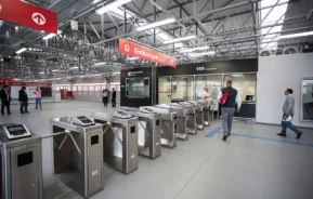Uma estação de trem ou metrô espaçosa e moderna com catracas de metal, uma placa de embarque acima e várias pessoas caminhando ou em pé nas proximidades, sob iluminação e tetos em estilo industrial.