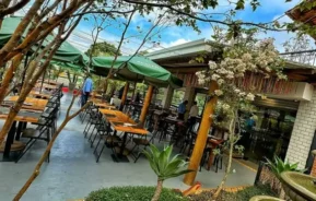 Pátio externo do restaurante com mesas e cadeiras de madeira sob guarda-sóis verdes, cercado por árvores e plantas. Uma pequena fonte e um lago estão em primeiro plano - perfeitos para apreciar o charme dos restaurantes em Embu das Artes.