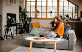Imagem de uma mulher de cabelos cacheados sentada em um sofá cinza em uma sala de estar moderna e aconchegante, falando ao telefone e segurando uma caneca. Grandes janelas, plantas e um espaço de trabalho são visíveis ao fundo