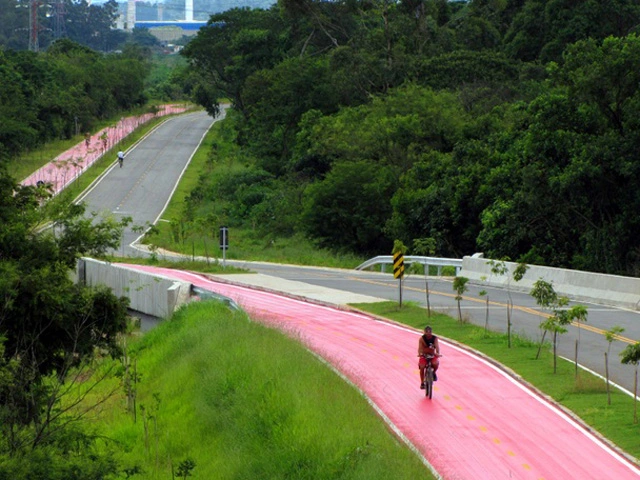 Imagem de uma pessoa andando de bicicleta em uma ciclovia rosa brilhante, cercada de vegetação, paralela a uma estrada comum, com outro ciclista à distância, no PET, em SP. Árvores e grama se alinham em ambos os lados das pistas.