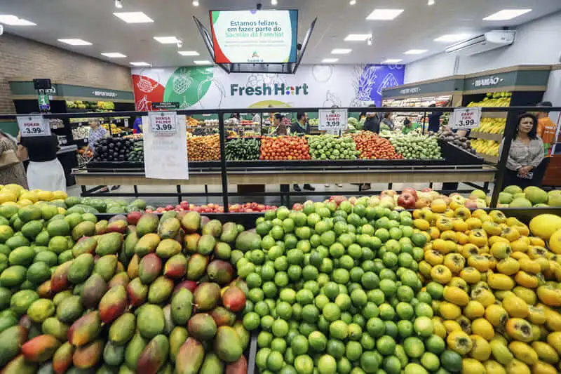 Imagem de uma seção de produtos de uma loja do Mercado Municipal de Curitiba mostra frutas bem organizadas, como mangas e maçãs. As placas exibem os preços e um grande logotipo do FreshMart pode ser visto ao fundo, acima das prateleiras de produtos. Os compradores estão navegando pela área
