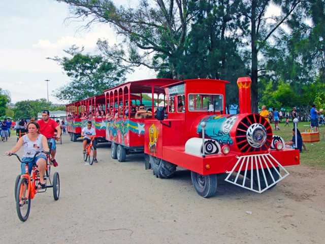 Imagem de um trem sem trilhos vermelho brilhante com desenhos coloridos transportando passageiros no Parque Ecológico do Tietê. Pessoas andam de bicicleta laranja nas proximidades, e árvores e outros visitantes são visíveis ao fundo