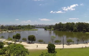 Imagem da vista do Parque Ecológico do Tietê, em São Paulo, em um dia ensolarado, mostra lago cercado por árvores e trilhas para caminhada. As pessoas passeiam e relaxam ao longo do caminho, com grama verde e um horizonte distante da cidade sob um céu azul com nuvens dispersas