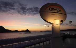 Um grande letreiro iluminado do Olimpo Restaurante fica à beira-mar ao pôr do sol, com um céu colorido, colinas distantes e uma passarela coberta que leva à água ao fundo.