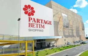 Um grande shopping center com uma fachada branca exibindo o logotipo vermelho e cinza e o texto Partage Betim Shopping. Há uma rampa amarela e uma estrada em frente, com um céu azul claro acima.