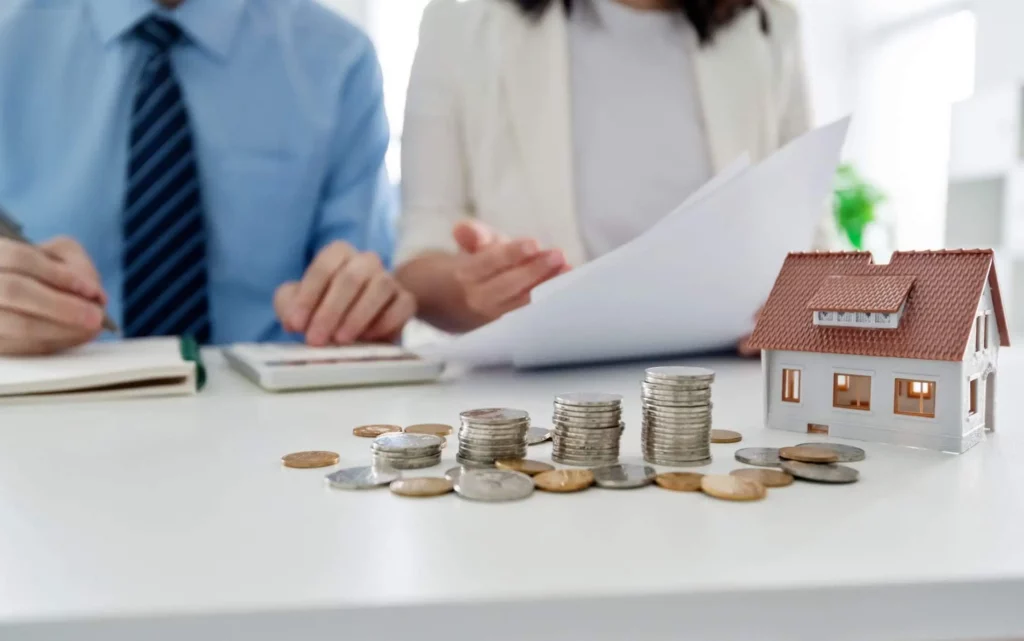 Imagem de duas pessoas sentadas em uma mesa com papéis, uma calculadora e uma casa modelo, com pilhas de moedas em primeiro plano