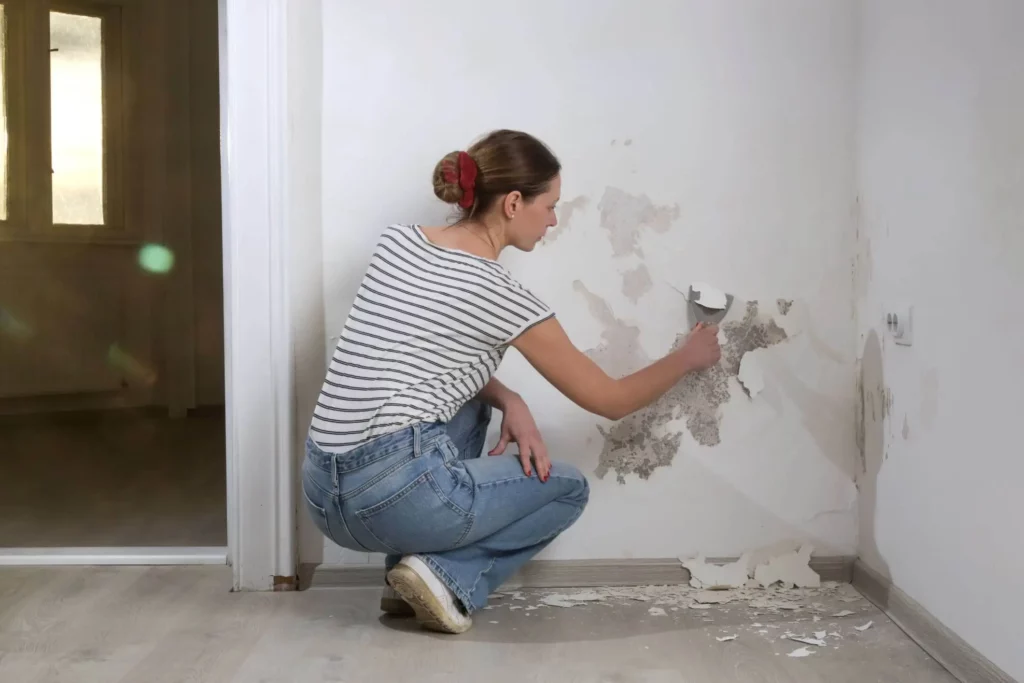 Imagem de uma mulher vestindo jeans e camisa listrada agachada ao lado de uma parede, usando um raspador para remover tinta descascada da superfície de uma parede branca em uma sala