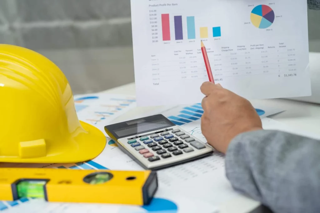 Imagem de uma pessoa apontando para um gráfico financeiro com um lápis. Nas proximidades, há um capacete amarelo, uma calculadora e um nível de bolha