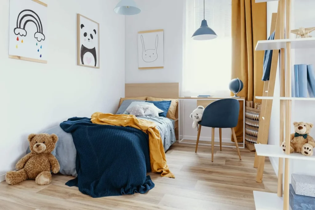 Imagem de um quarto infantil aconchegante com uma cama coberta com cobertores azuis e amarelos, um ursinho de pelúcia no chão, uma escrivaninha com uma cadeira e um globo terrestre, arte de animais na parede e cortinas amarelo-mostarda em uma janela clara.