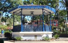 Um gazebo de ferro azul com grades decorativas e luminárias fica em um parque ensolarado, cercado por plantas e árvores. Prédios coloridos podem ser vistos ao fundo.
