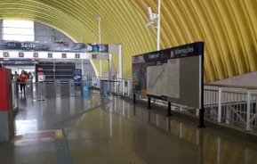 Interior de uma moderna estação de metrô com teto curvo amarelo, barreiras de bilhetes, quadros de informações e algumas pessoas caminhando perto dos sinais de saída. O piso é brilhante e reflete os arredores.