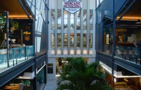 Vista interna do Mercado Municipal de Niterói, com uma palmeira central, arquitetura moderna com grades de vidro, lojas em dois andares e luz natural passando por janelas altas.