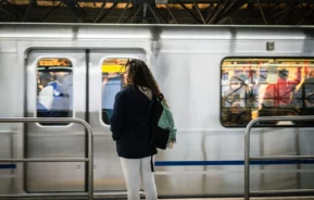 Uma pessoa com longos cabelos cacheados, vestindo uma jaqueta escura e calça clara, está em uma plataforma de metrô com uma mochila enquanto um trem passa ao fundo.