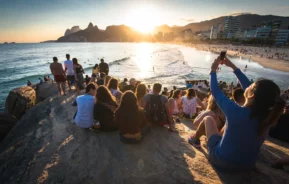 Um grande grupo de pessoas está sentado em rochas à beira-mar, assistindo ao pôr do sol em uma praia. Alguns estão tirando fotos com seus telefones. A cena inclui ondas, surfistas e prédios da cidade ao fundo. A cena acontece no Arpoador, no Rio de Janeiro, para muitos o pôr do sol mais bonito do Brasil.