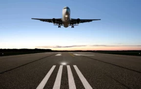 Imagem de um avião comercial pousando em uma pista ao pôr do sol, com céu limpo ao fundo e marcações da pista visíveis em primeiro plano