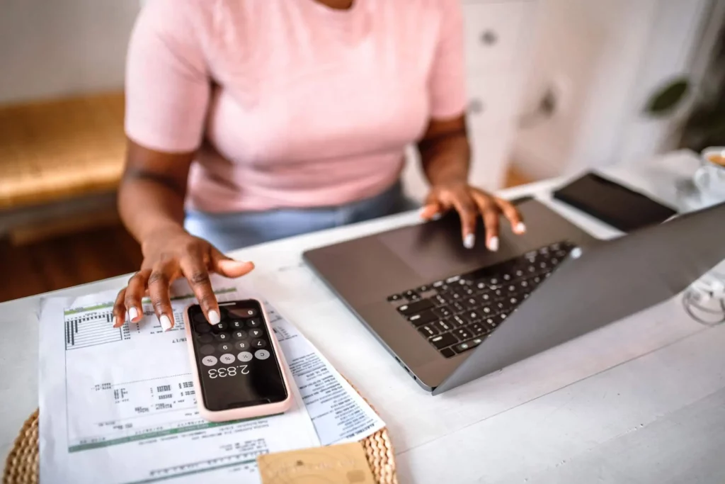 Imagem de uma pessoa vestindo uma camisa rosa usa um laptop e uma calculadora de smartphone enquanto analisa documentos financeiros em uma mesa.