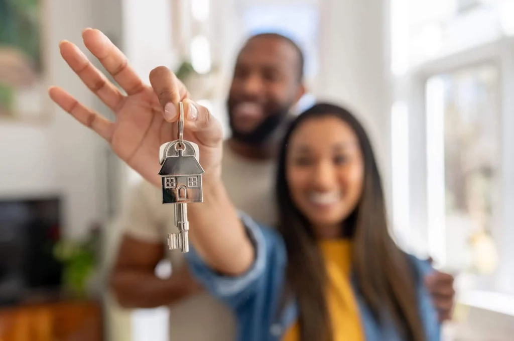 Imagem de um casal sorridente em um ambiente interno, com a mulher segurando um chaveiro em forma de casa e uma chave em direção à câmera, simbolizando a compra de uma nova casa. O fundo é claro e está fora de foco