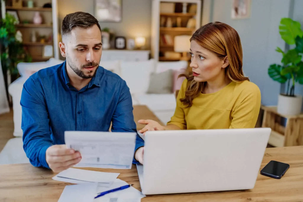 Imagem de um homem e uma mulher sentados à mesa com um laptop e papéis, parecendo preocupados e envolvidos em uma conversa séria em um ambiente doméstico aconchegante.