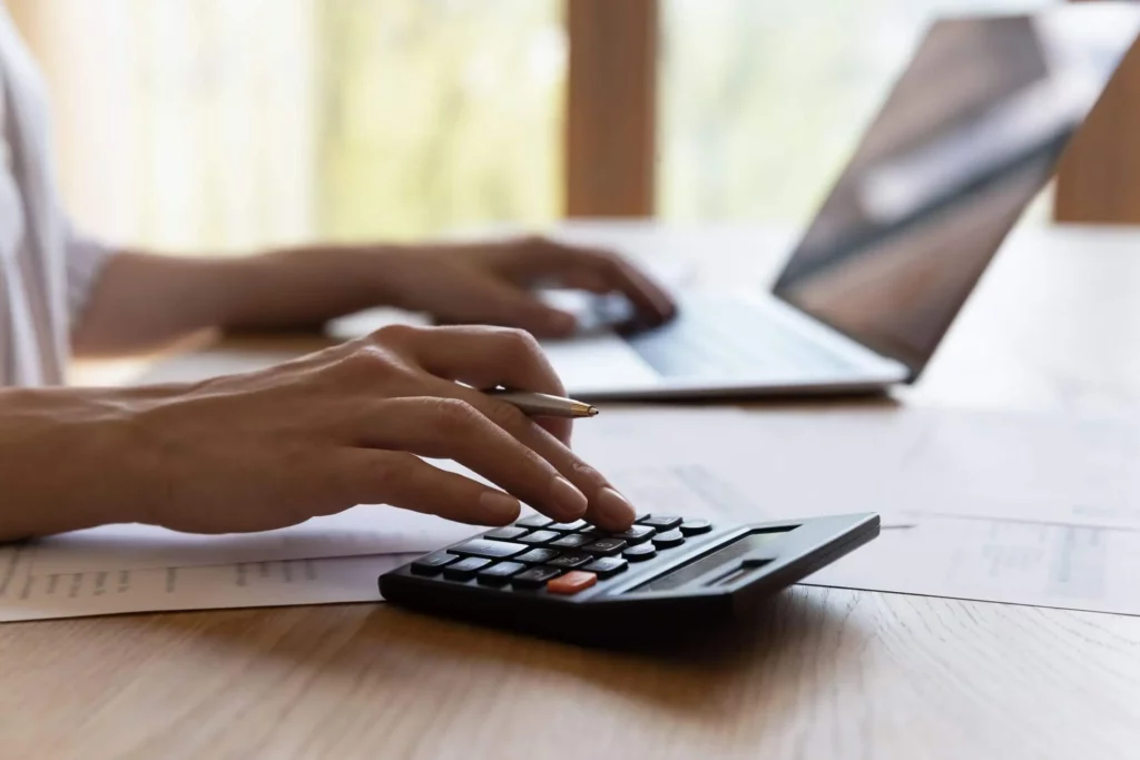 Imagem de uma pessoa usando uma calculadora e digite em um laptop em uma mesa de madeira, com papéis espalhados