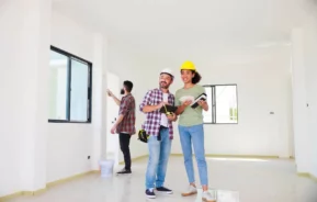 Imagem de dois trabalhadores da construção civil, um homem e uma mulher, estão sorrindo em uma sala iluminada e inacabada, segurando planos e cadernos. Outro trabalhador, ao fundo, verifica uma parede. Todos vestem roupas casuais; a mulher usa um capacete amarelo.
