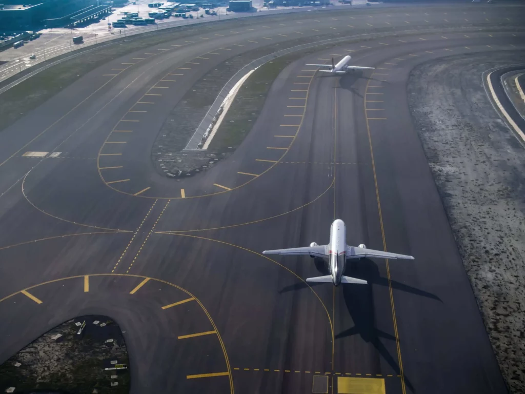 Imagem de dois aviões comerciais em pistas paralelas em um aeroporto, cercados por marcações amarelas e pistas de taxiamento que se cruzam, com edifícios e veículos do aeroporto visíveis ao fundo