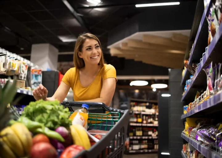 Imagem de uma mulher de camisa amarela sorrindo enquanto empurra um carrinho de compras cheio de frutas e legumes frescos por um corredor em um supermercado.