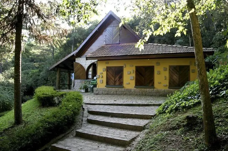 Imagem de Casa da Bruxa, no Bosque do Alemão, em Curitiba, mostra um chalé charmoso com paredes amarelas, portas de madeira e um telhado inclinado que fica entre uma vegetação exuberante e árvores, com degraus de pedra que levam à entrada. A luz do sol é filtrada pelas folhas.