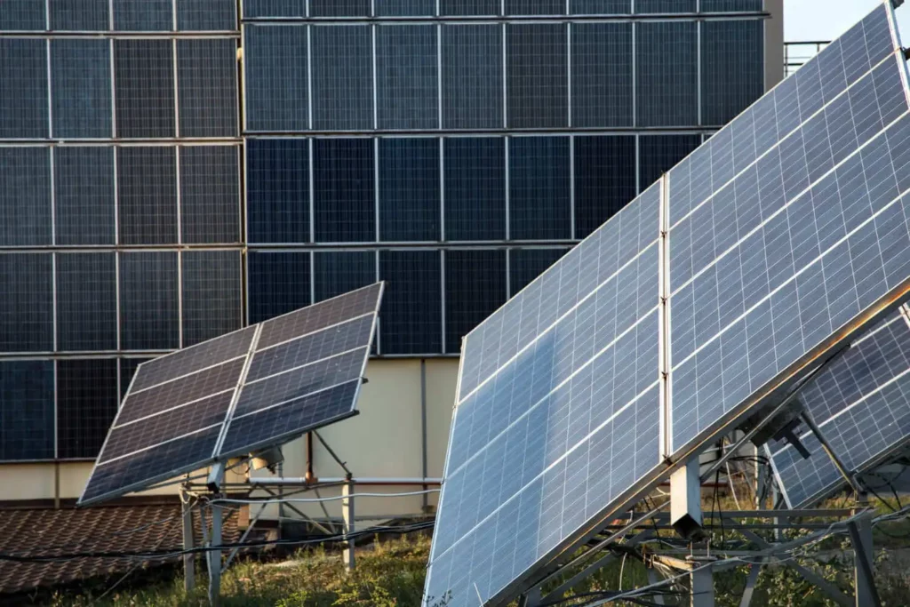 Imagem de vários painéis solares montados ao ar livre em estruturas metálicas, inclinados em direção ao sol, com mais painéis solares instalados verticalmente em um edifício ao fundo. A cena sugere um foco em energia renovável.