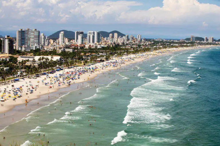 Imagem da vista aérea do mar, prédios, vegetação e banhistas na faixa de areia da Praia da Enseada, no Guarujá