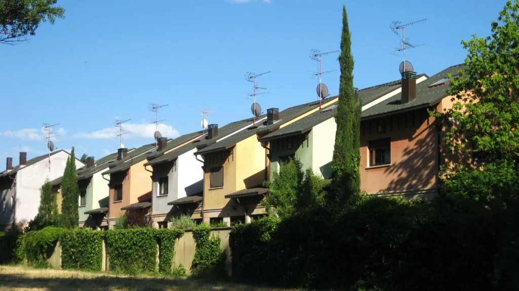 Imagem de uma fileira de casas geminadas coloridas com telhados inclinados, antenas parabólicas e varandas, ladeadas por arbustos verdes e árvores sob um céu azul claro.
