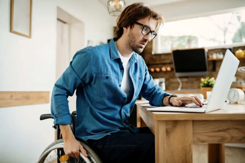 Imagem de um homem de óculos e camisa jeans sentado em uma cadeira de rodas em uma mesa de madeira, trabalhando em um laptop em uma cozinha moderna e bem iluminada.