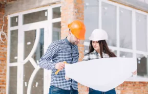 Duas pessoas usando capacetes e camisas xadrez estão do lado de fora de um prédio em construção, olhando juntas para uma grande planta. A estrutura atrás deles tem tijolos expostos e janelas grandes.